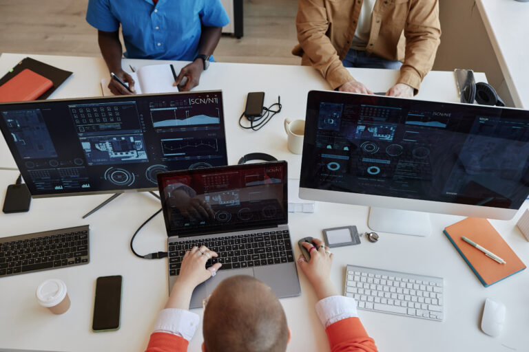 Above view of three intercultural coworkers analyzing cyber data on screens of their computers while sitting by large table in office